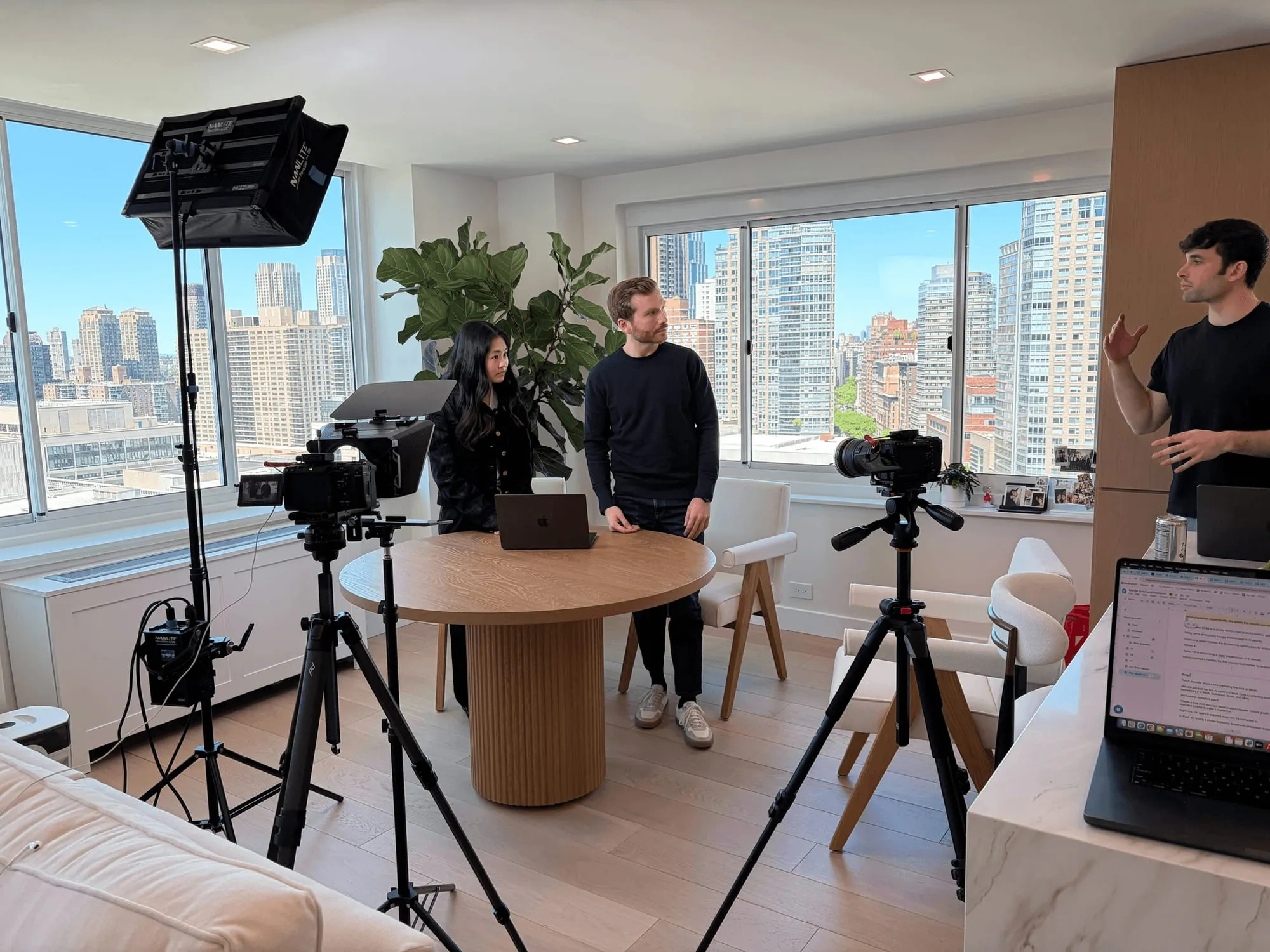 Behind the scenes of a shoot: two cameras and a teleprompter pointed at three people seated at a round table, with a city skyline through the windows.