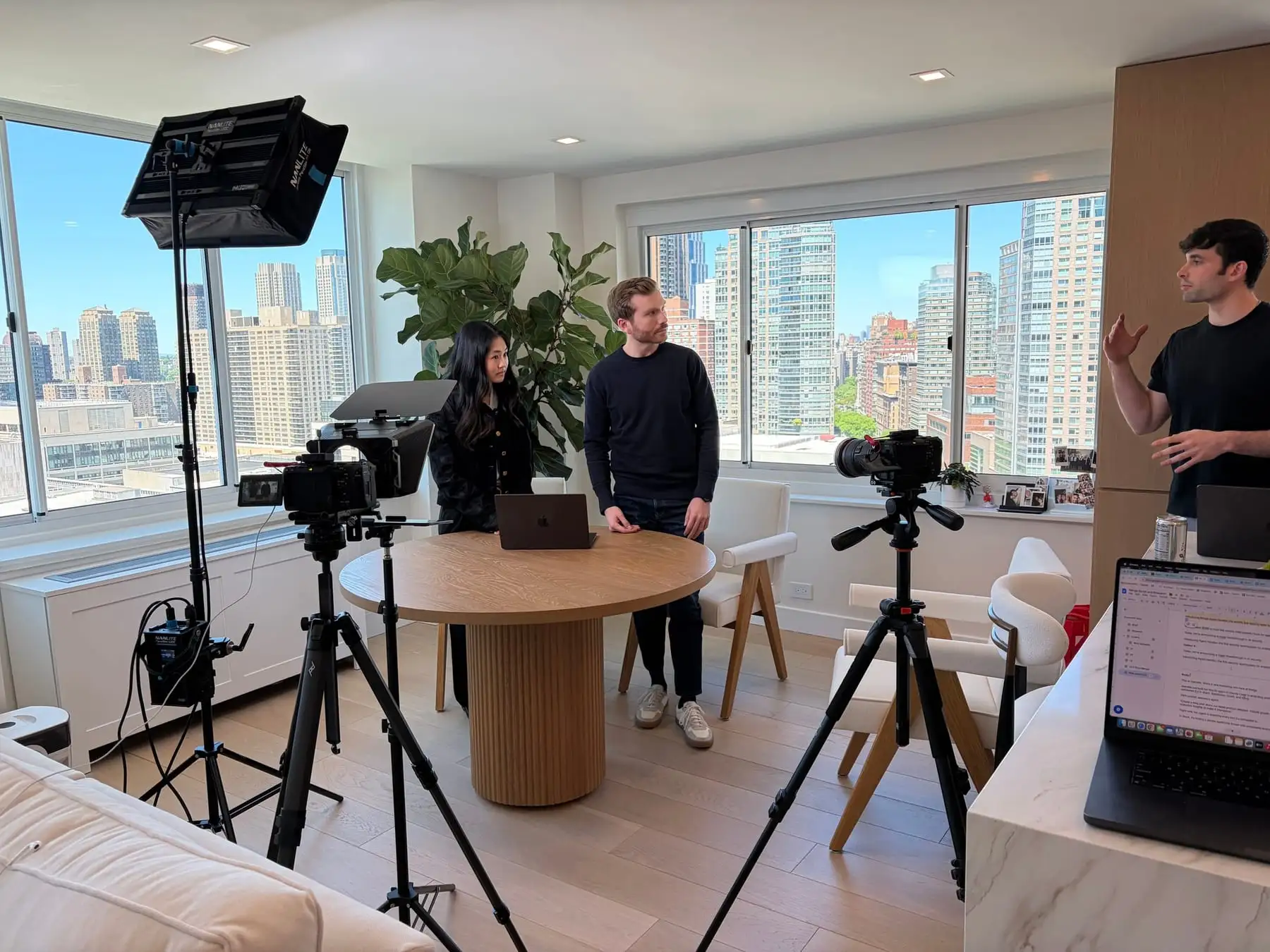 A shoot in a high-rise apartment: two cameras on tripods and a light around a round table, with three people talking through the shot.