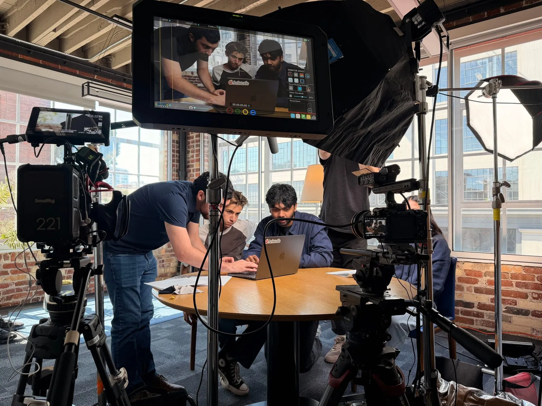 On set: a monitor in the foreground shows the shot while three people work at a laptop on a round table, surrounded by cameras and lights.