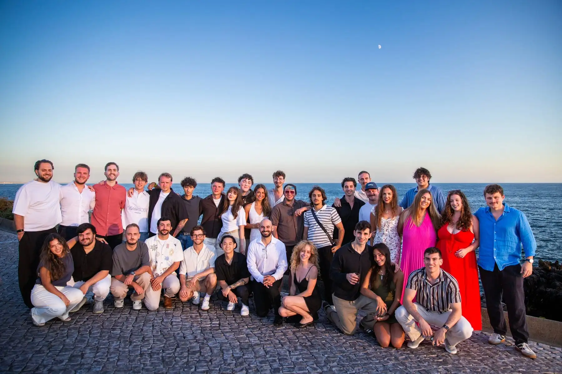 The team, standing and kneeling in two rows for a group photo on a cobbled seafront at dusk.